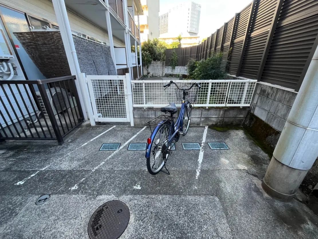 青い自転車が停まっているコンクリート製の駐輪場、背後には壁と植栽が見える静かな住宅街の一角