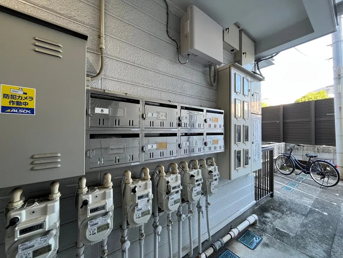 - 外観の郵便受けと電気メーター、セキュリティカメラの警告サイン、隣に自転車が停まるエントランスの様子