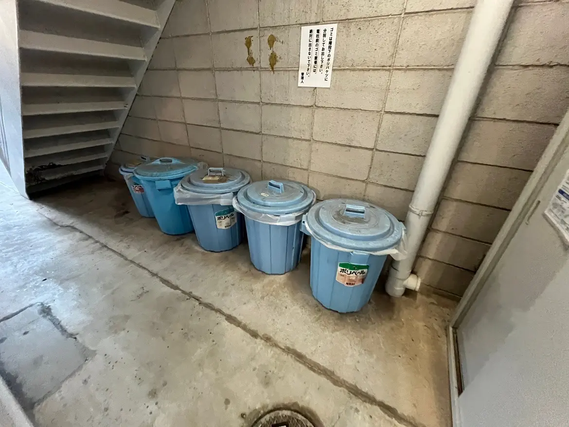 - 地下の廊下に並べられた青いゴミ箱、壁には掲示物があり、階段の隣に設置された状態