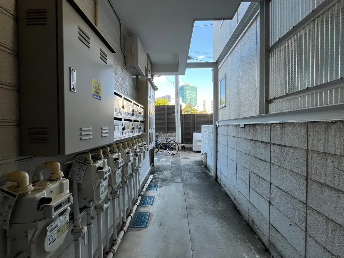- 建物の外側に並ぶガスメーターと郵便受け、奥に自転車が見える幅の狭い通路の風景