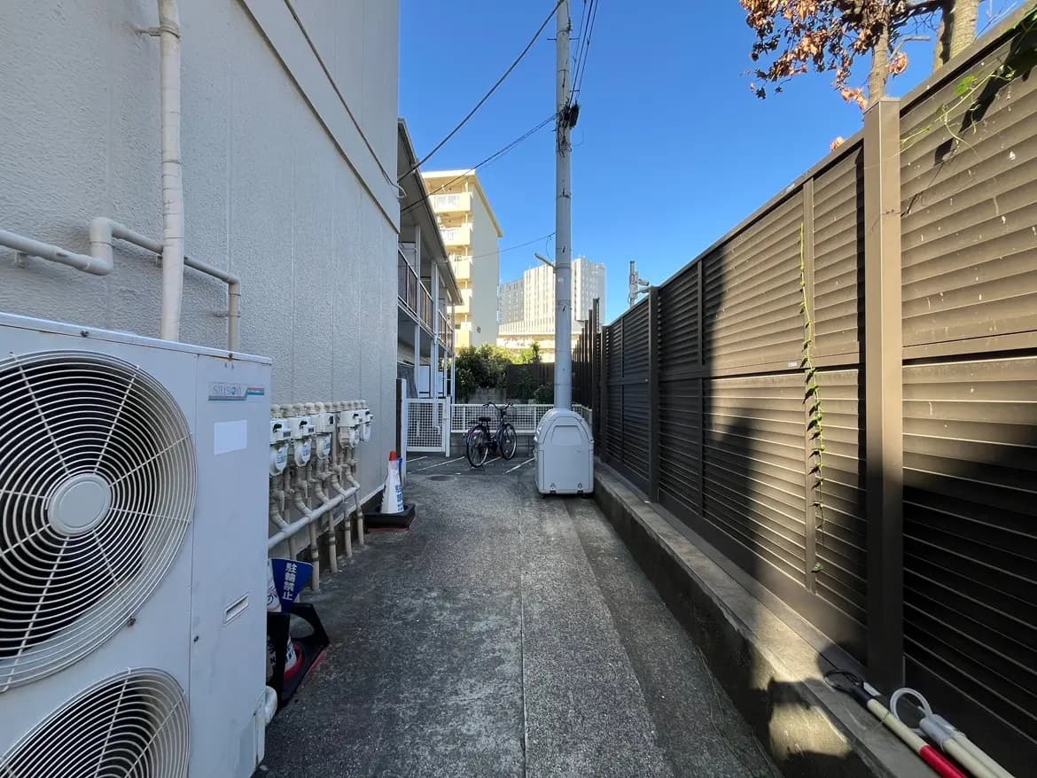 - 建物の側面に設置されたエアコンとガスメーターが並んでいる狭い通路、青空の下の住宅街の風景