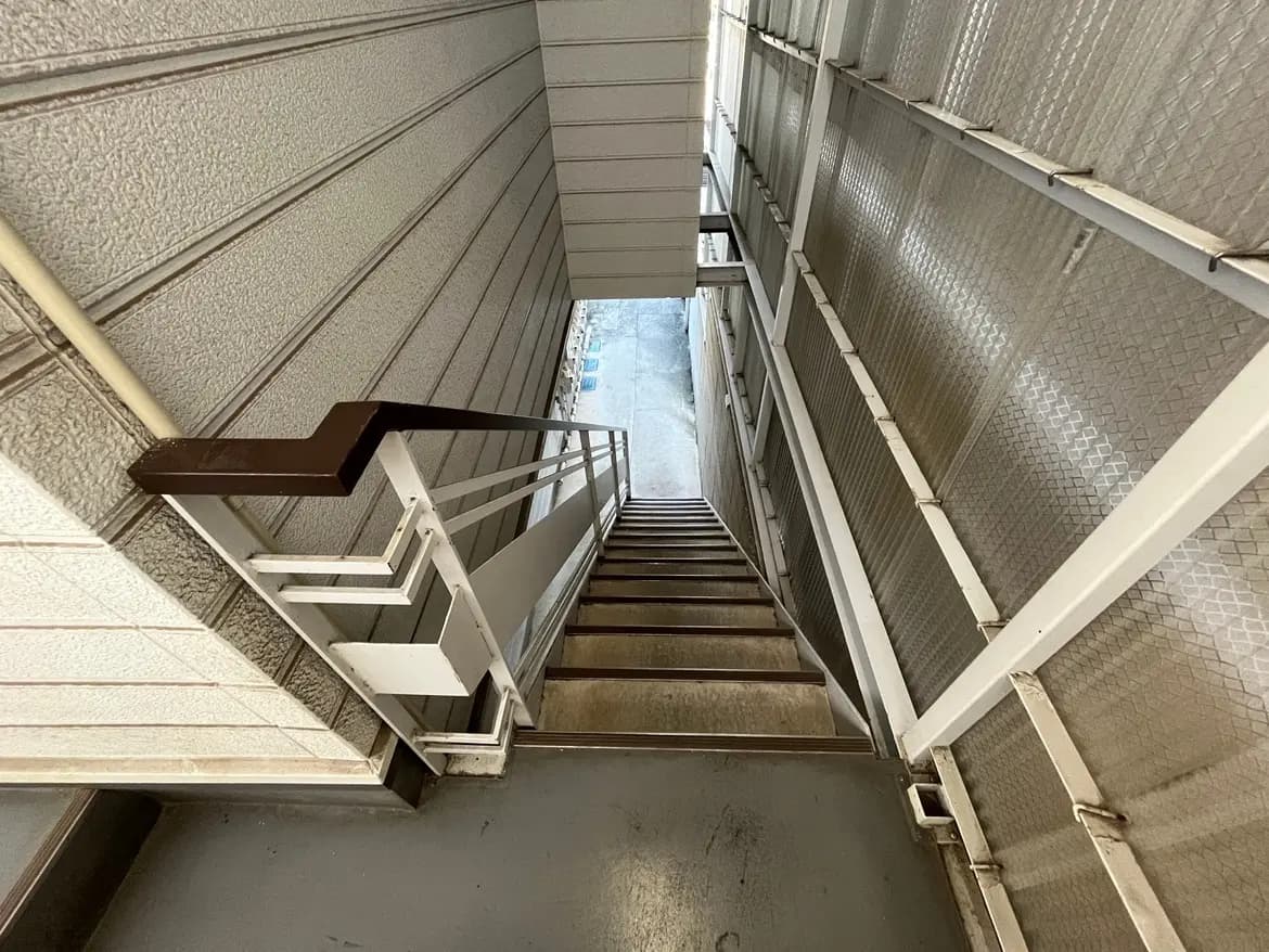 - 鉄製の手すりがある階段の上から見下ろした視点、光が差し込む外の景色が見える廊下の写真