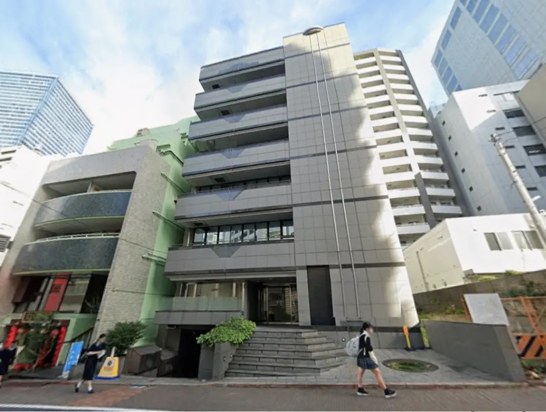 - 都市部に位置する現代的な建物の外観、グレーの外壁と階段、周囲に高層ビルが立ち並ぶ風景