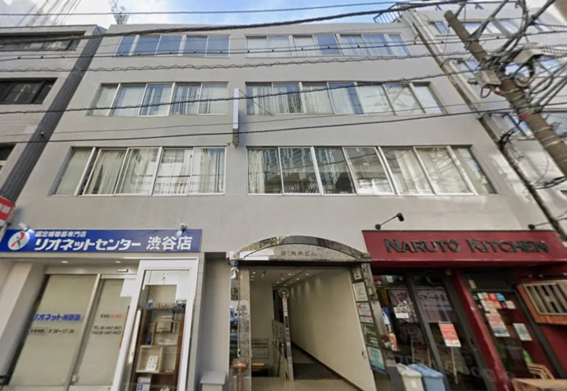 - 渋谷にあるリオネットセンターの外観、近隣にあるナルトキッチンの店舗と明るい日差しが差し込むビル正面の写真