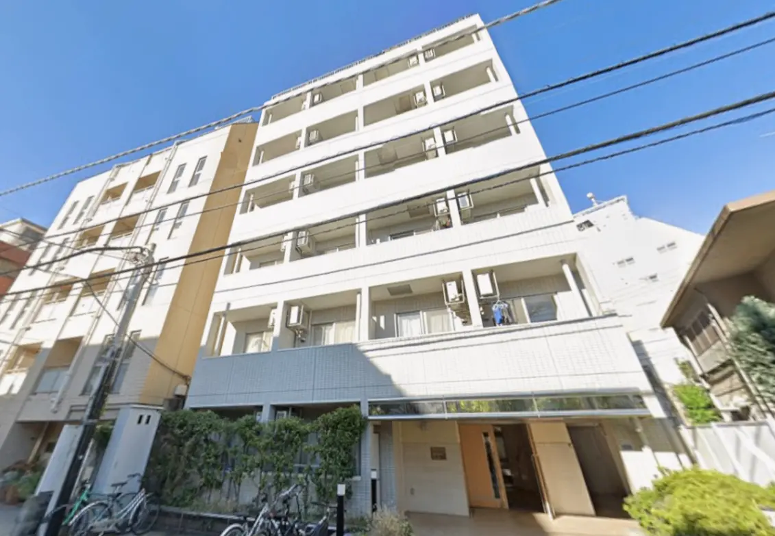 - ブロック状の白い外観を持つ5階建てアパート、バルコニーにはエアコンが設置され、近隣に自転車が置かれたエントランスの様子