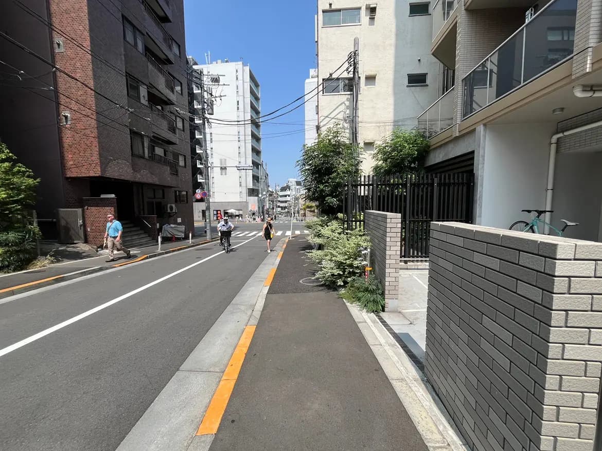 - 都市の街並みを描写した画像、歩行者と自転車が通る道、周囲に高層ビルと緑が見える晴れた日中の風景