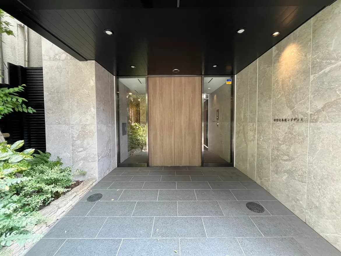 - モダンな建築デザインのエントランス、木製の扉と石材の壁、緑の植栽が施された明るい入り口の風景