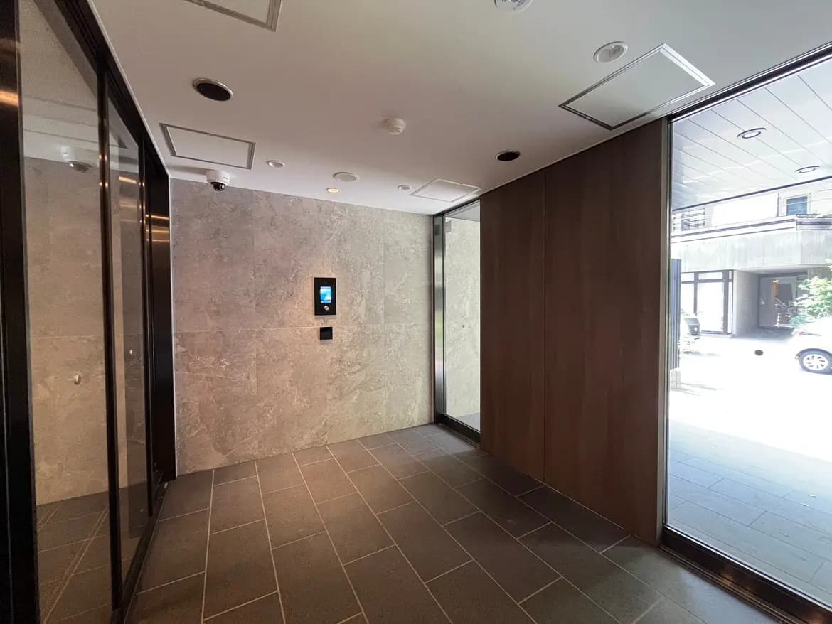 - モダンなエントランスホール、グレーの石材と木材の壁、ガラスのドアから自然光が入る空間、セキュリティモニター付き