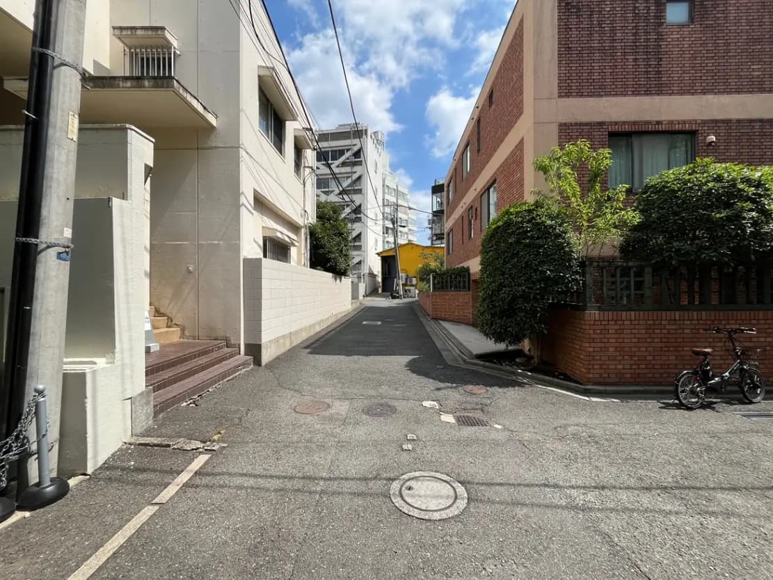 - 明るい青空の下、住宅街の狭い道路と周囲の建物、並ぶ緑の生垣と自転車が置かれた風景