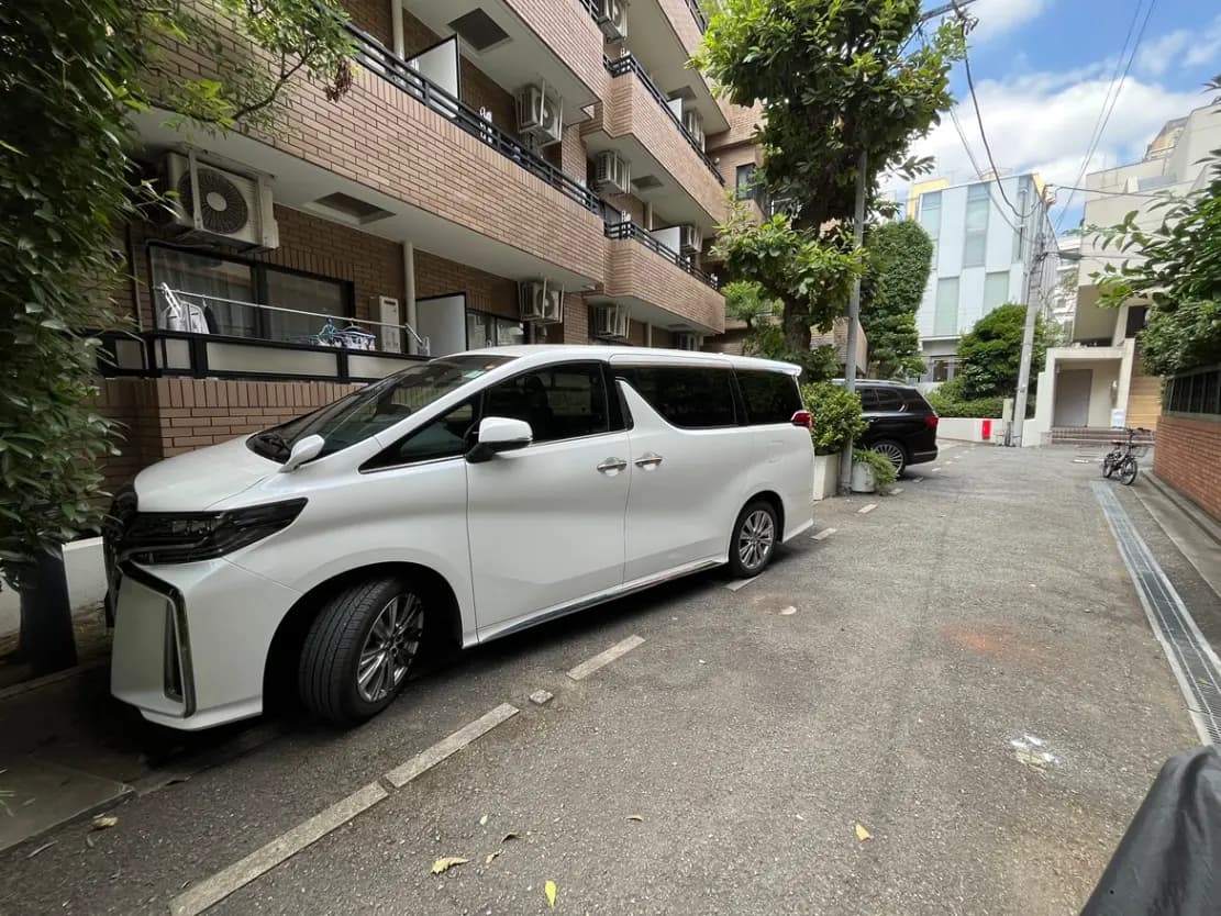 - 白いミニバンが静かな住宅街の路上に停車している様子、周囲にはマンションと緑が広がる風景
