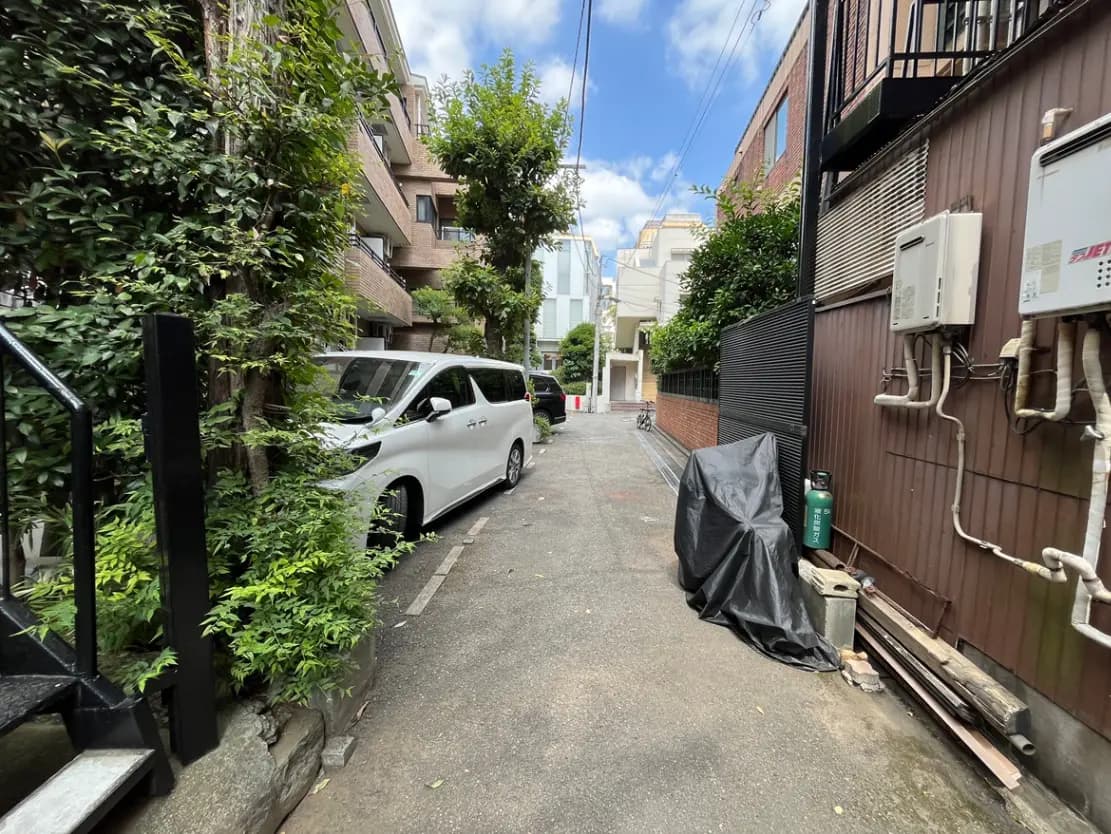 - 緑に囲まれた狭い路地、白い車が停まっており、周囲には住宅と物置が見える風景
