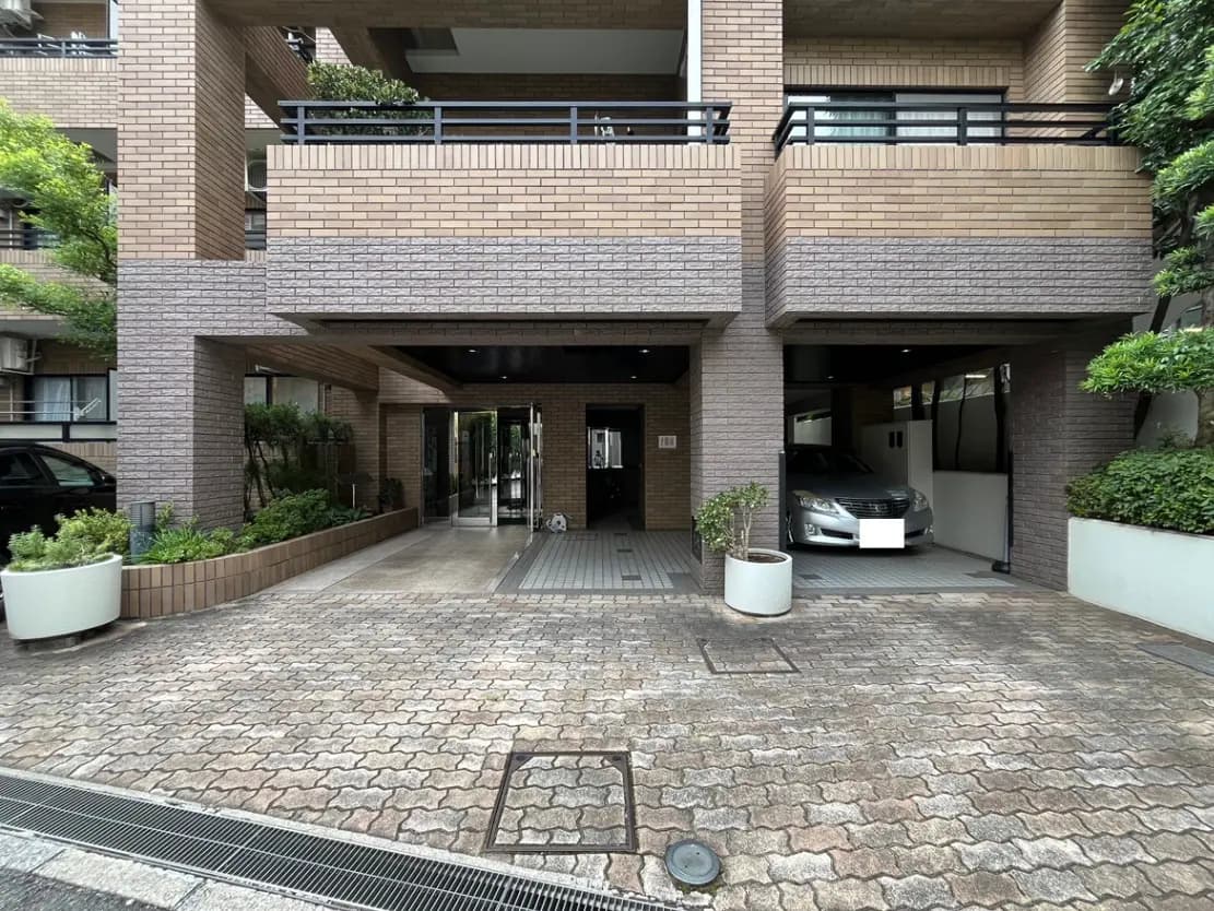 - モダンなマンションのエントランス、レンガ造りの外観と緑の植栽、駐車スペースがある風景