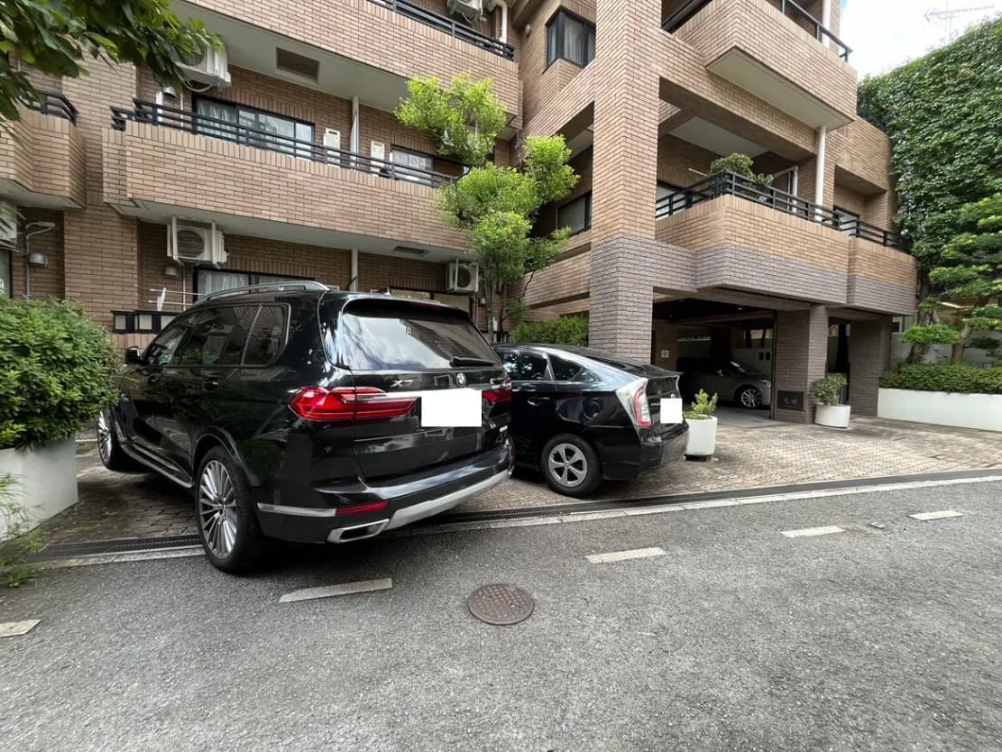 - 高級マンションのエントランス前に駐車された黒いSUVとセダン、周囲には緑の植栽がある風景