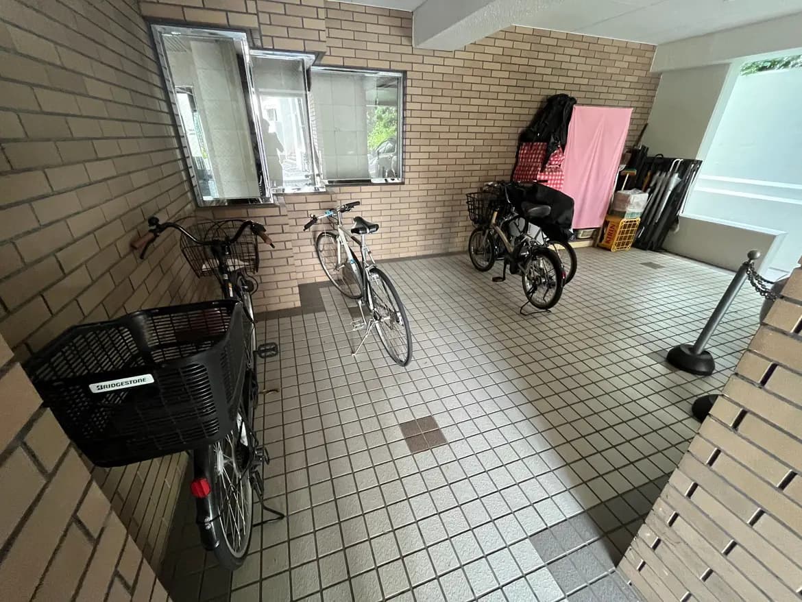 - 自転車置き場の内部、レンガ調の壁とタイルの床、複数の自転車が停められている様子、窓からの自然光が差し込む空間