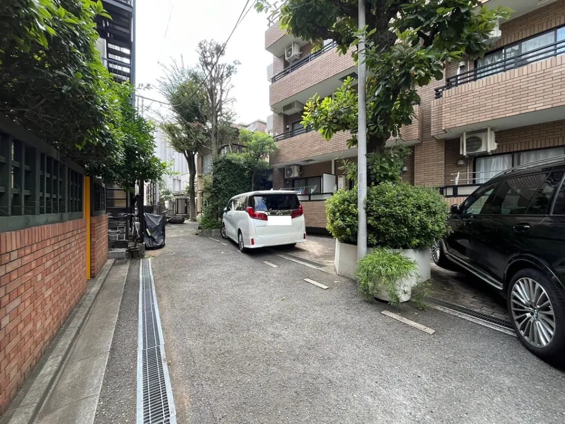 - 落ち着いた雰囲気の住宅街の駐車場、白い車と黒い車、緑の木々とレンガ壁が見える風景