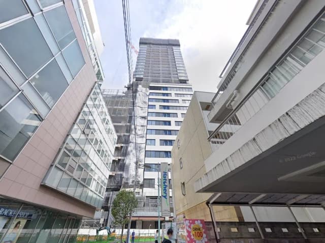高層ビルの建設現場、足場やネットがかかる様子、近代的な商業施設が周囲にある都市景観