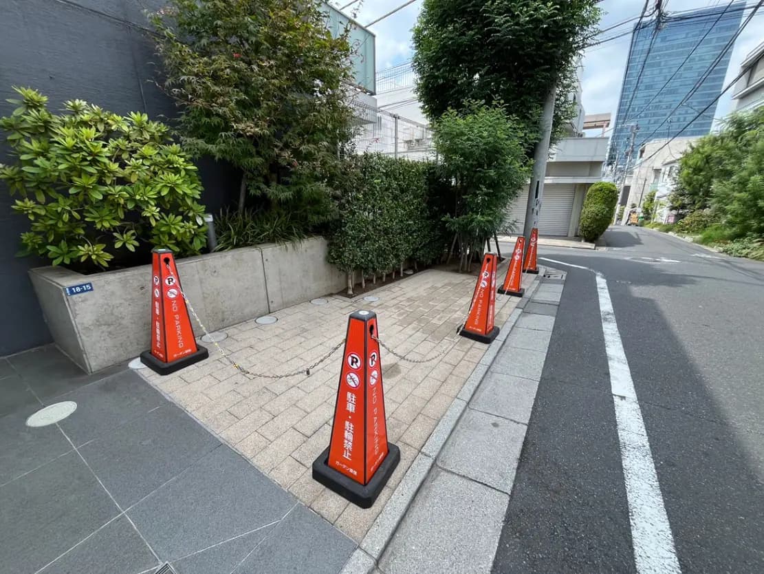 - 道路脇に設置された「駐車禁止」のオレンジ色のコーンと緑の植栽、舗装された歩道の風景