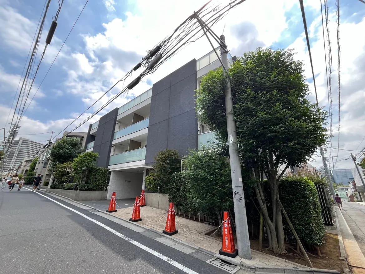 - モダンなデザインのマンション外観、青空と白い雲が広がる背景、周囲には緑豊かな木々と交通標識がある風景