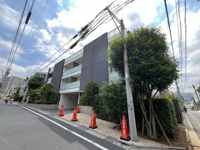- モダンなデザインのマンション外観、青空と白い雲が広がる背景、周囲には緑豊かな木々と交通標識がある風景