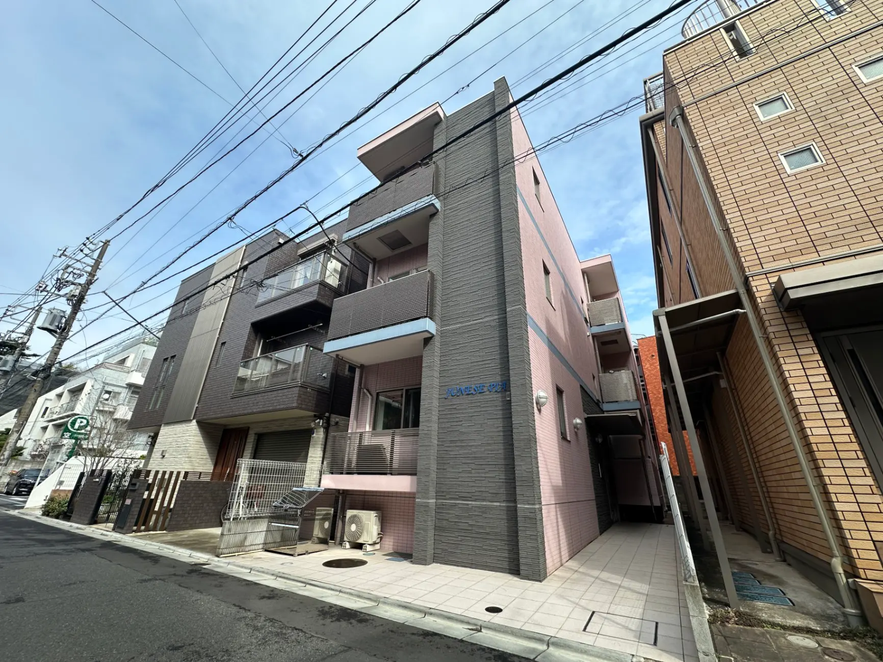 都市の住宅街にあるピンク色の3階建てアパート、近隣にモダンな建物と駐車場、晴天の下での街並み