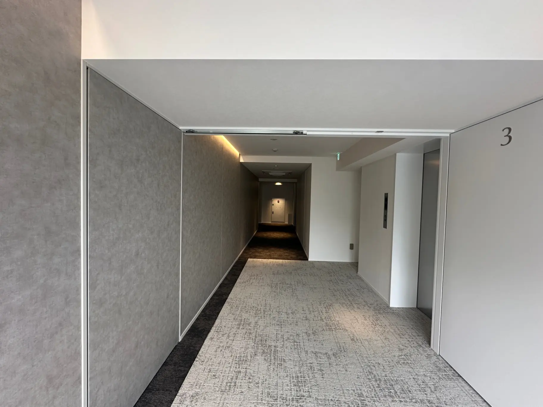 シンプルでモダンなデザインの廊下、グレーのカーペット敷き、壁には部屋番号表示、照明が足元を照らすインテリア