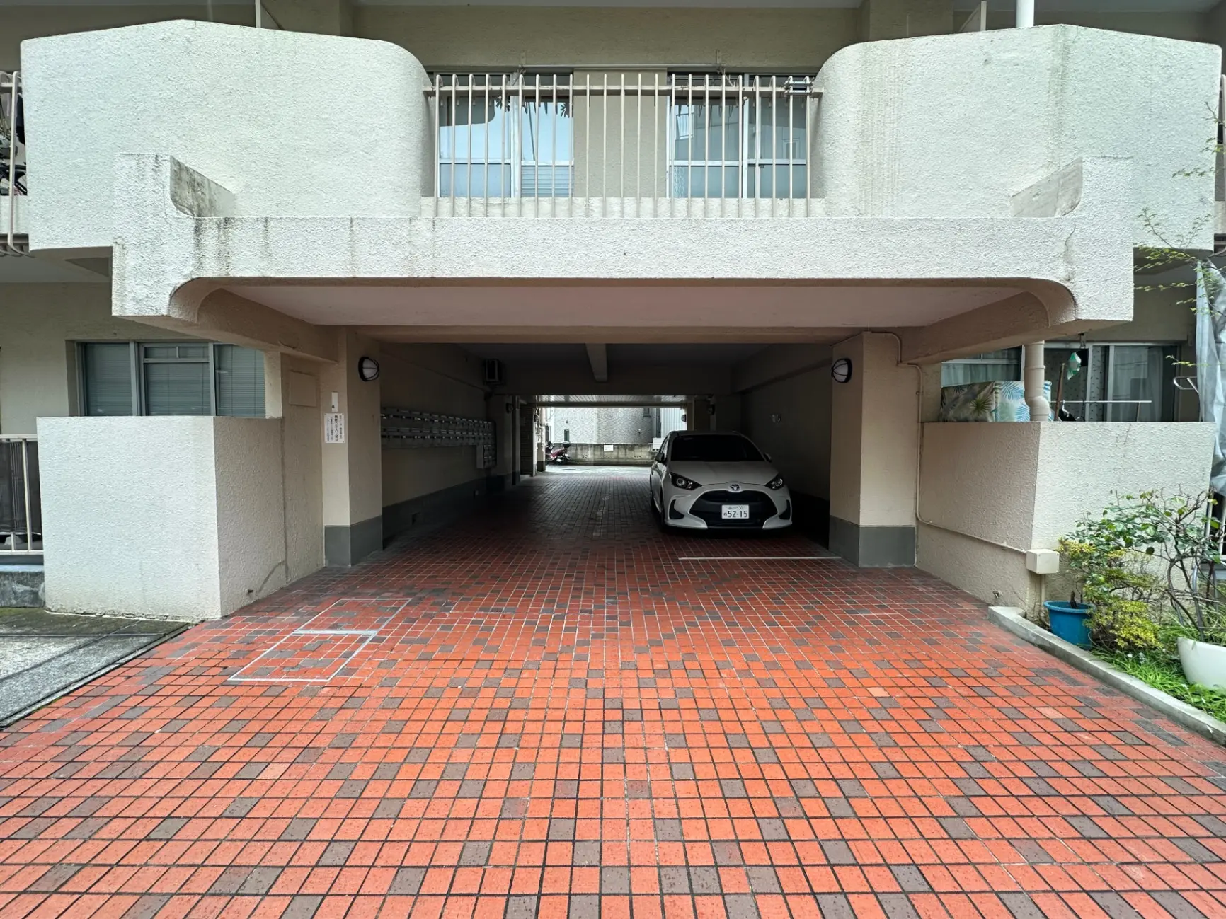 都市型マンションの駐車場、白い外壁、赤色タイルの床、屋根付き駐車スペースに駐車中の白い乗用車