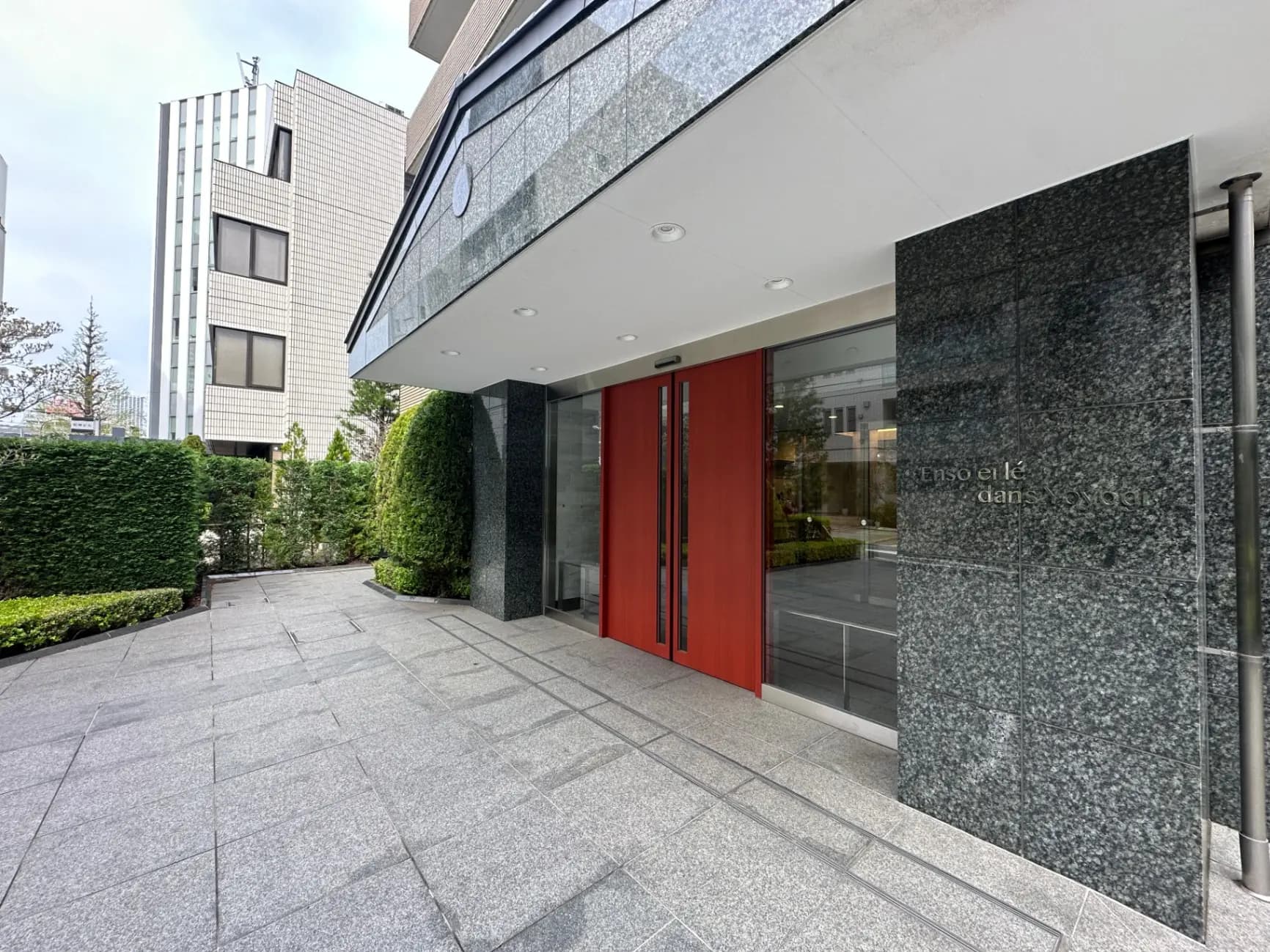 モダンでスタイリッシュなビルの入り口、緑豊かな植栽と赤いドアが目を引く外観デザイン