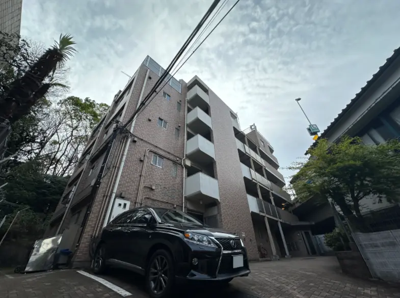 都会の住宅街にある現代的な外観のマンションと黒い車、青空と電線が見える街角