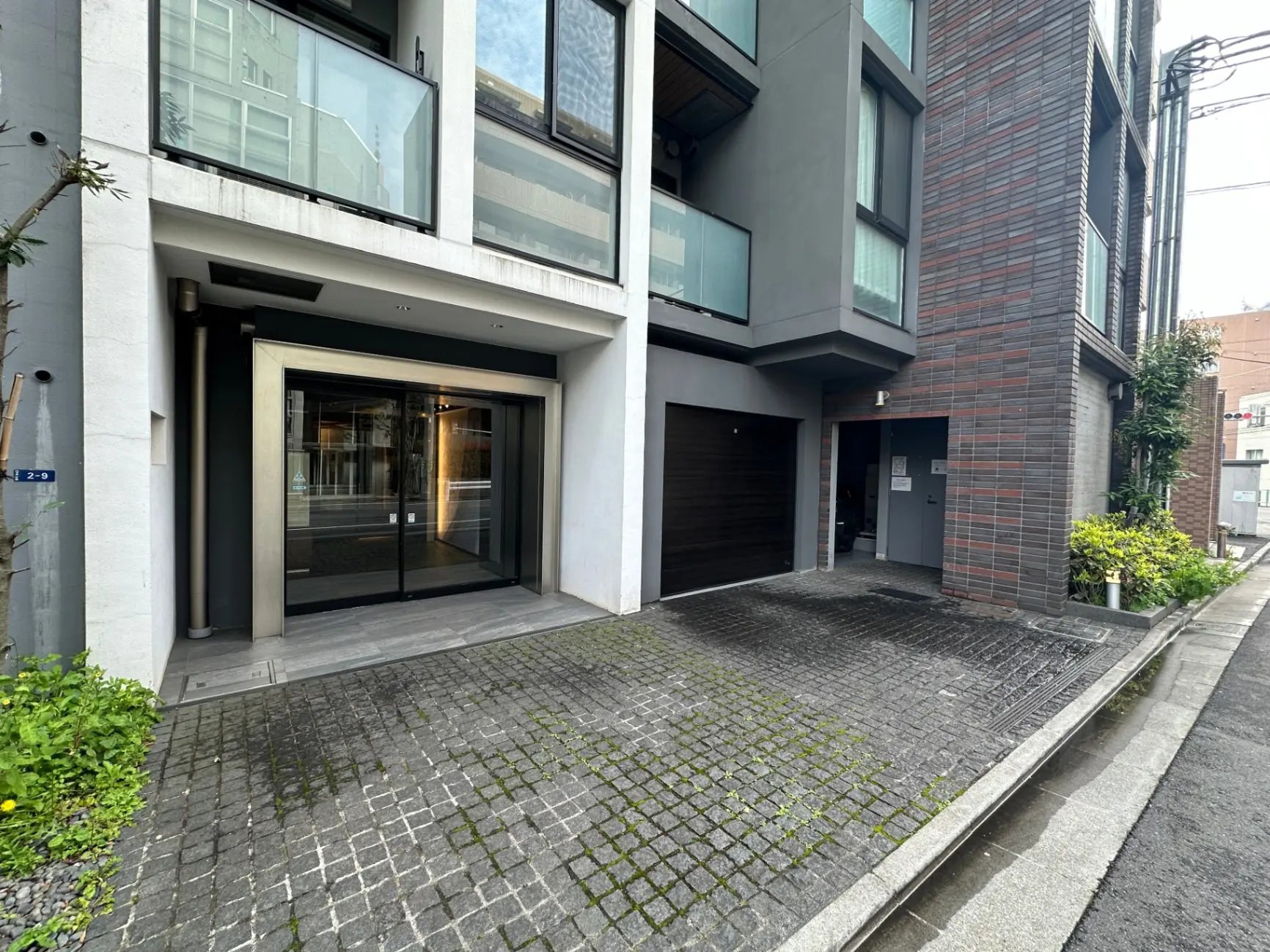 モダンなデザインの外観、グレーの壁、大きなガラス戸と緑の植栽が映える都市型アパートのエントランス