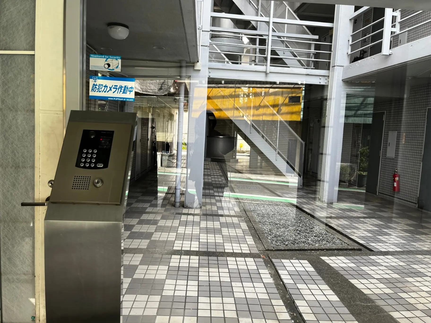 都市の住宅街にあるアパートのエントランスとインターホン、白黒のタイルが敷かれ、階段と防災用品が配置されている外観