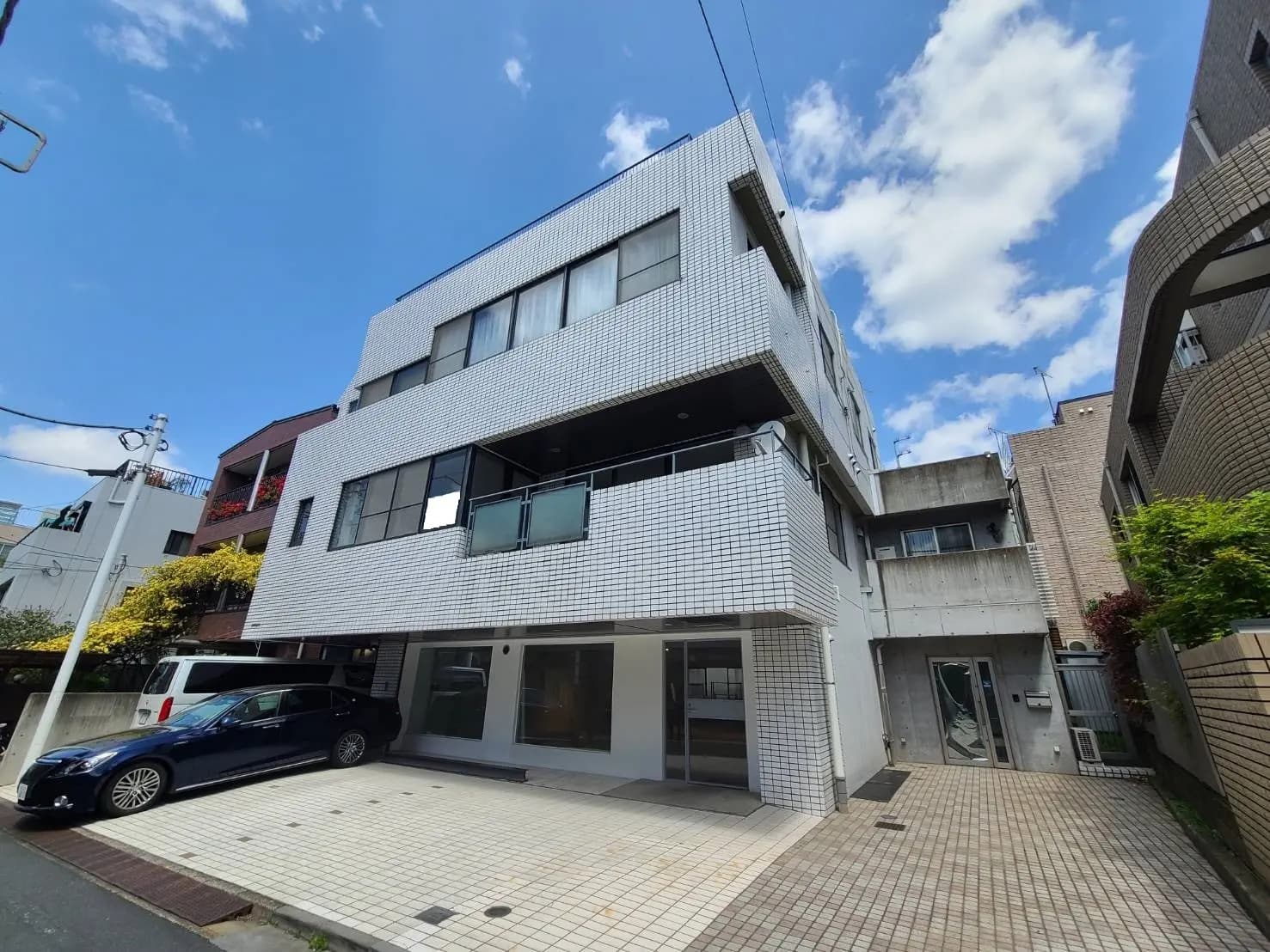 都会の住宅街にあるモダンな白いタイル張りの建物、駐車スペースとブルーの車、晴れた日の青空と雲