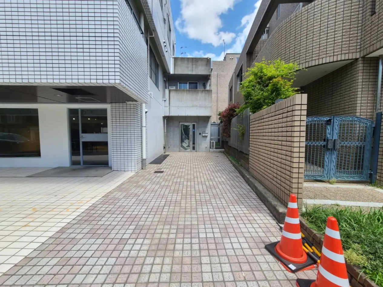 日中の都市風景、白い建物の入り口、赤と白の交通安全コーン、緑豊かな植栽がある歩道
