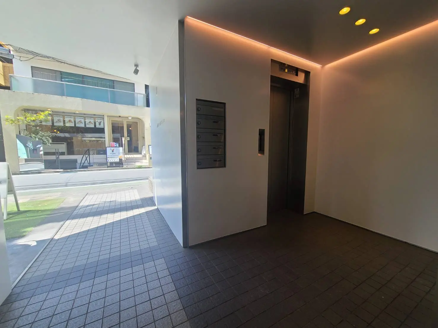 エントランスホールの開放的なデザイン、グレーのタイル床、LEDライトで照らされた天井、エレベーターと郵便受けがあるインテリア