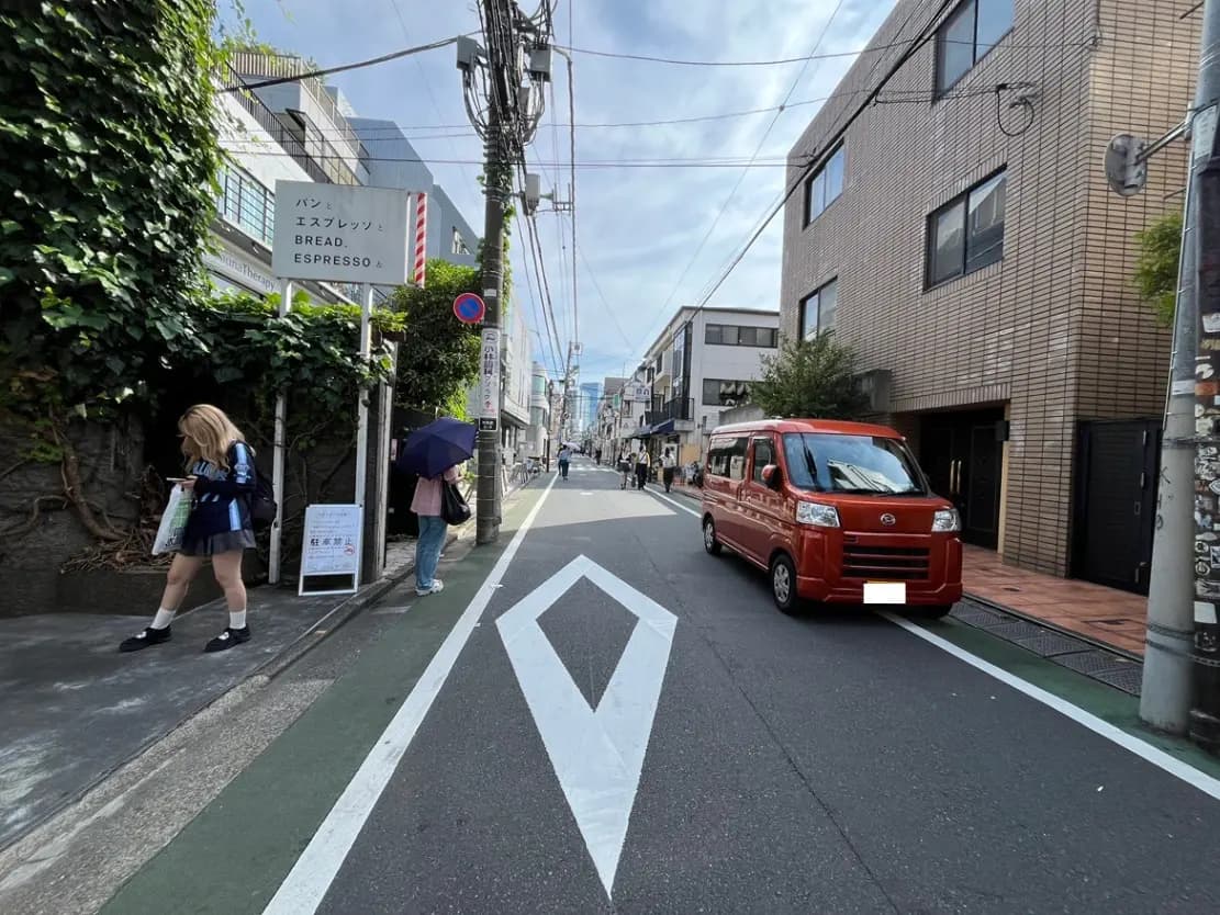 - 東京の街並みを背景にした通り、赤いミニバンと歩行者が見える風景、人気のパン屋の看板と緑の植生が特徴的なエリア