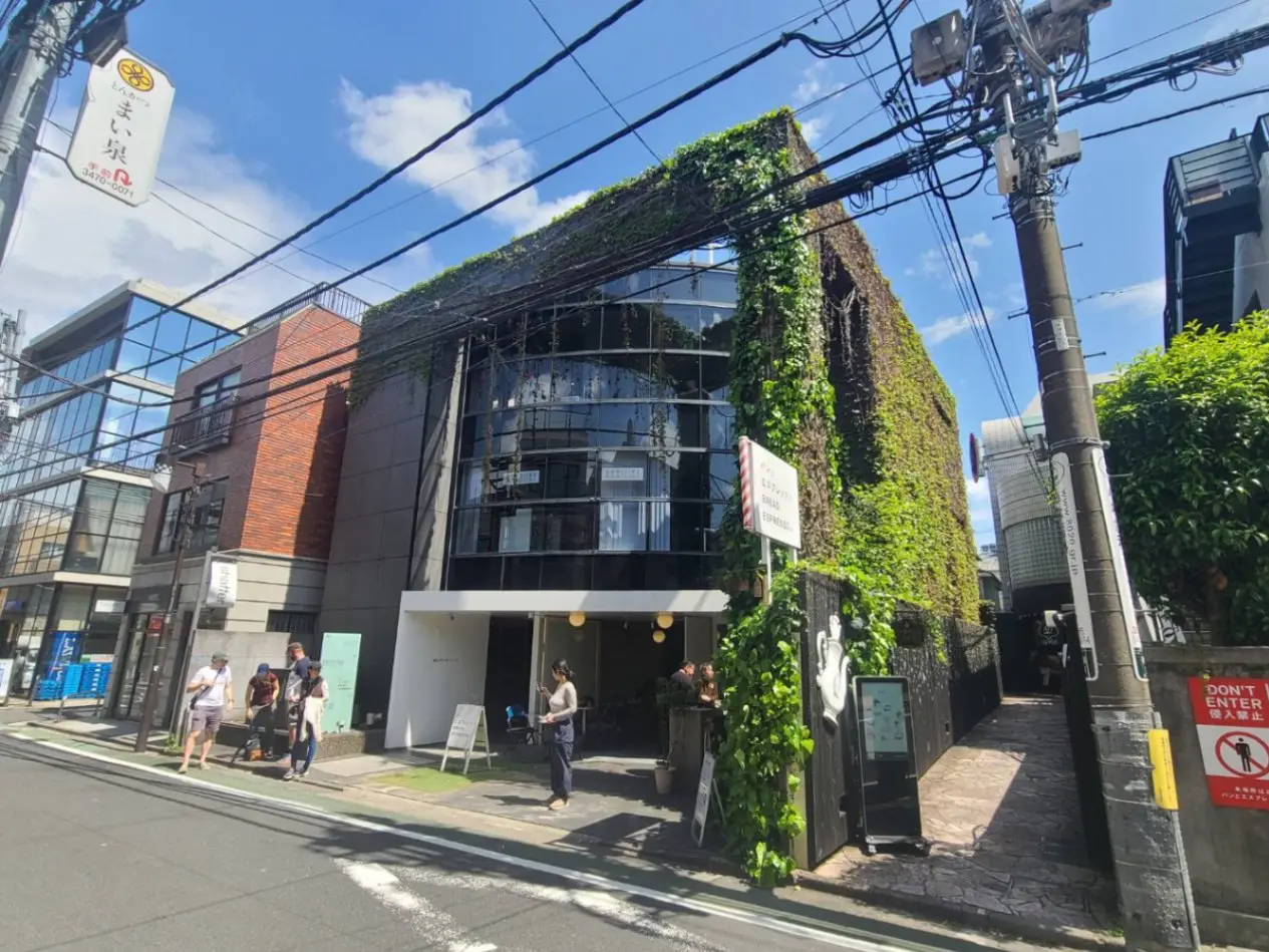 都会の中の緑豊かなビル、歩道を歩く人々、青空と電線が交差する景色