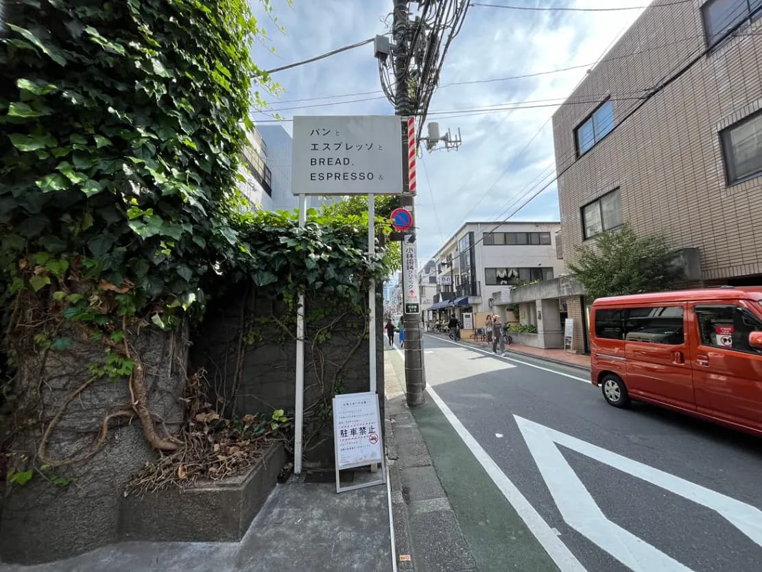 - 館の入口に立つ「パンとエスプレッソ」看板、周囲には生い茂る緑と都市の風景が広がる通りの風景