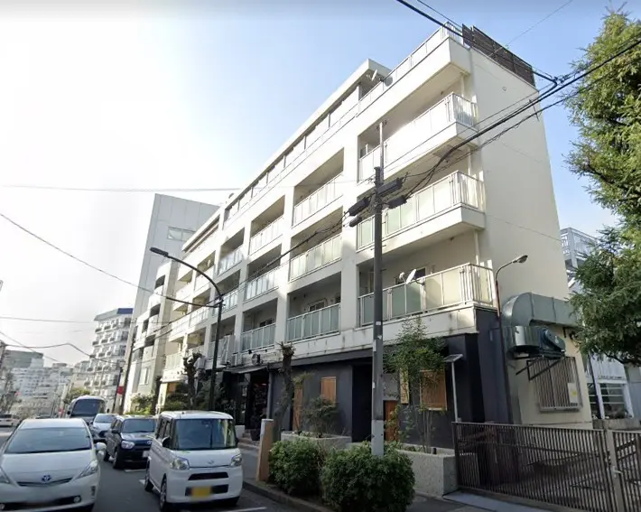 白い外壁とバルコニー付きのモダンなマンション、通り沿いに駐車された車と街路灯が見える外観
