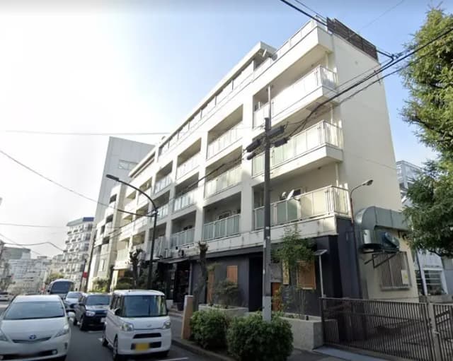 白い外壁とバルコニー付きのモダンなマンション、通り沿いに駐車された車と街路灯が見える外観