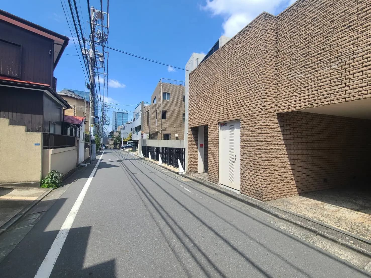 日本の静かな住宅街の路地、右手にレンガ造りのモダンな建物、左手に電柱と一軒家が並ぶ風景