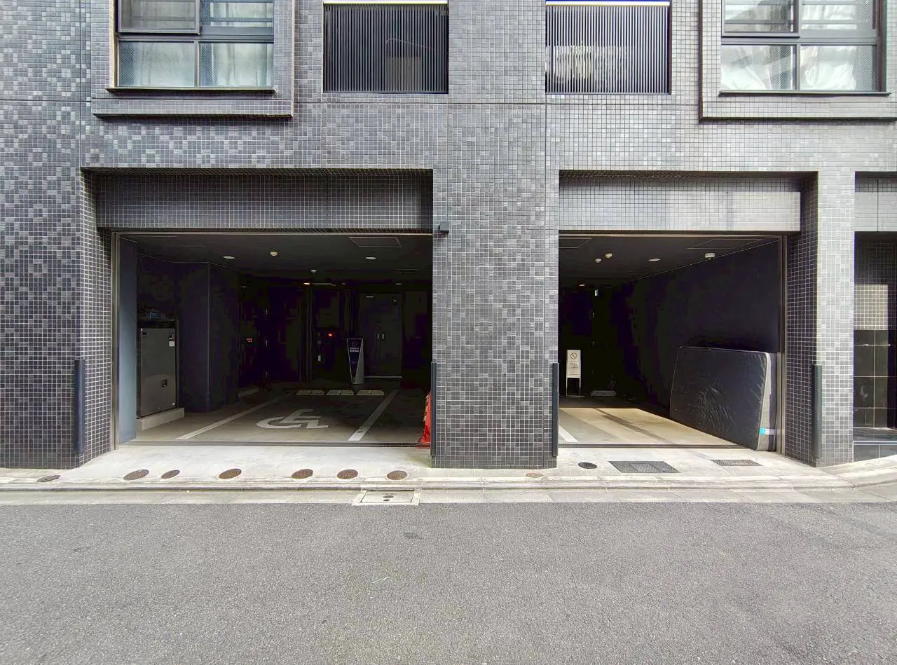 - モダンな外観のビル前にある2つの駐車スペース、車椅子マーク付きの駐車区画と暗いトンネルのような入り口が見える街角の風景