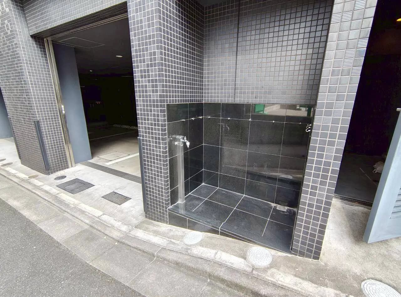 - 黒いタイルの外壁に囲まれたシャワースペース、周囲には舗装された歩道と交通用のマンホールが見えるエントランスの風景