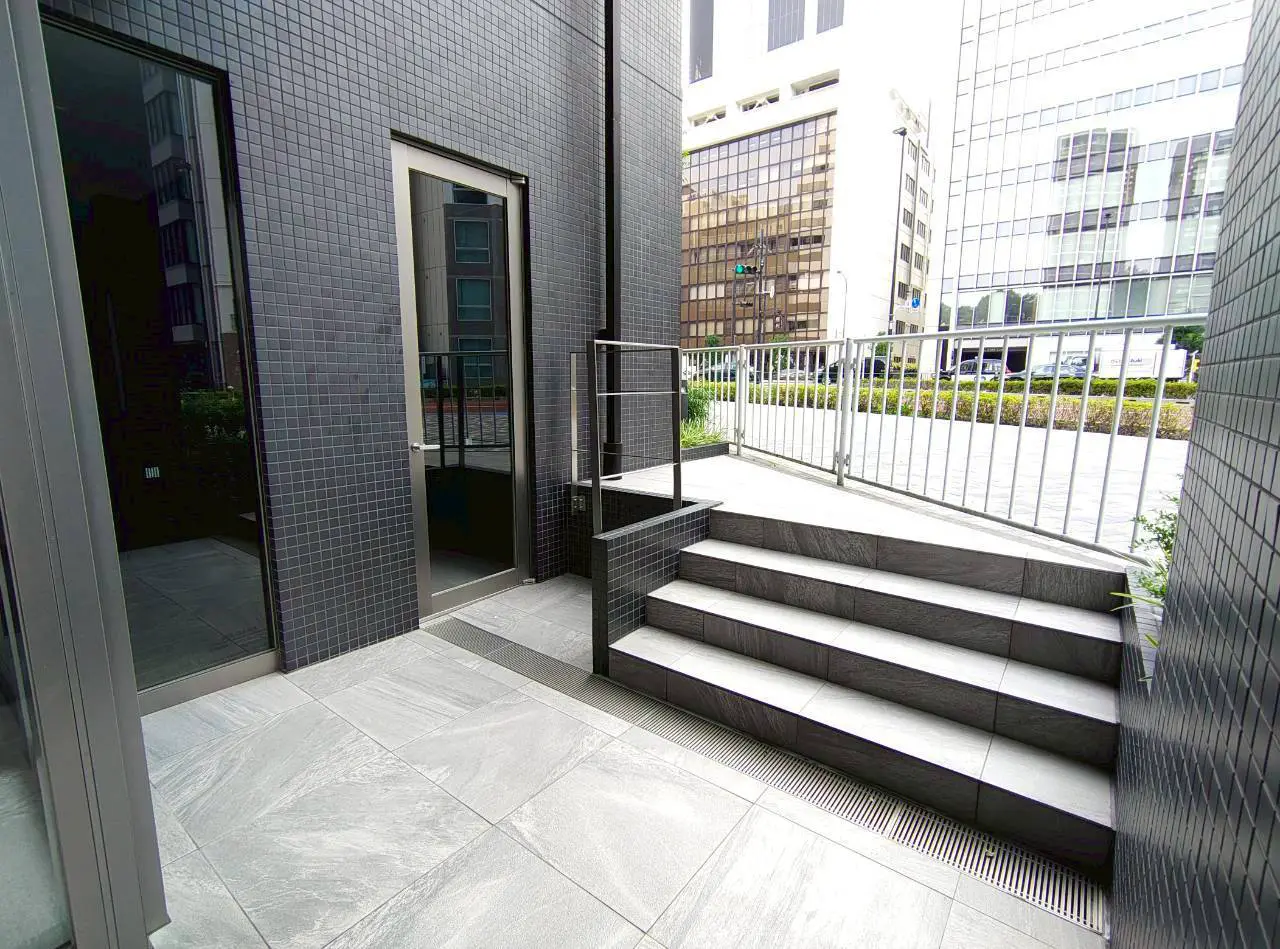 - モダンなデザインの外観、ステップと手すりがあるエントランス、周囲にビルが立ち並ぶ屋外スペース