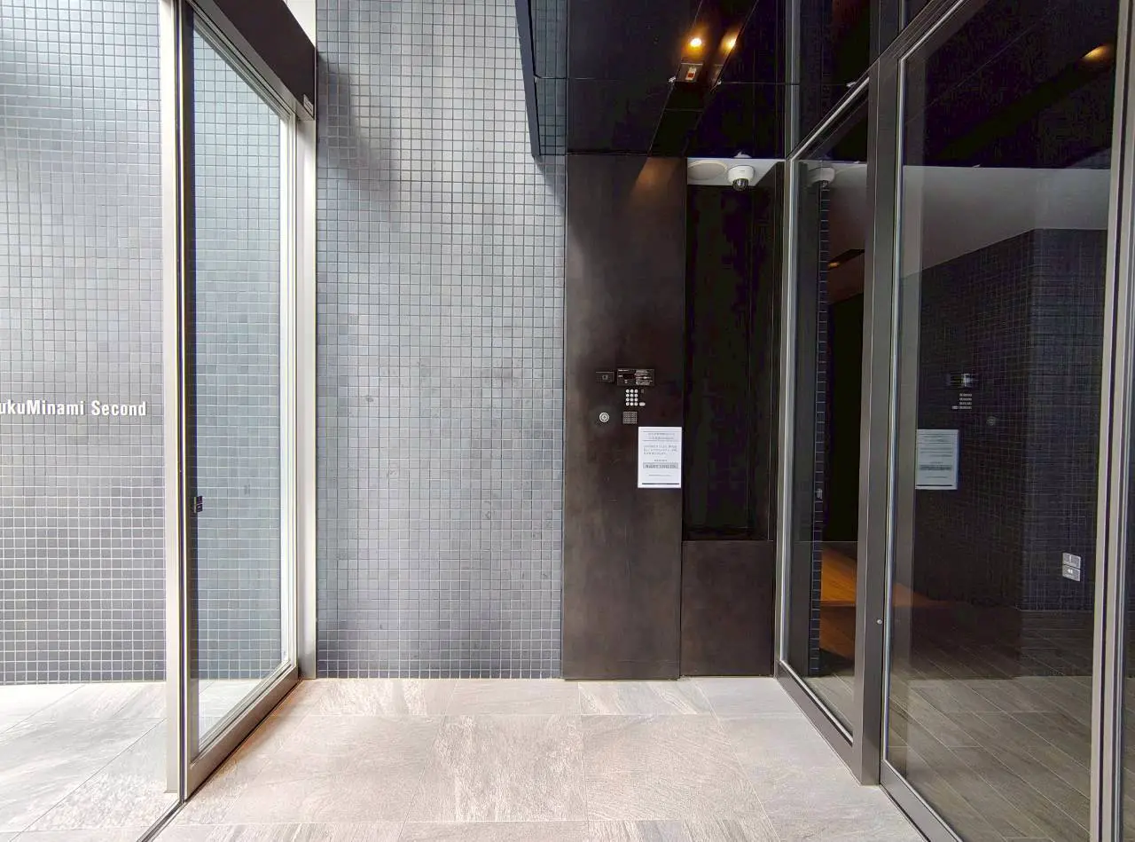 - モダンなエントランスの内部、グレーのタイル壁とガラスのドア、シンプルなデザインのインターフォンが設置された空間