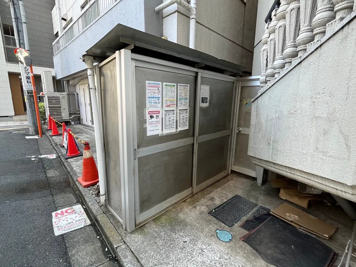 - 錆びた金属製のゴミ置き場と周囲のコーン、迷惑行為を禁止する掲示物が貼られた街角の風景