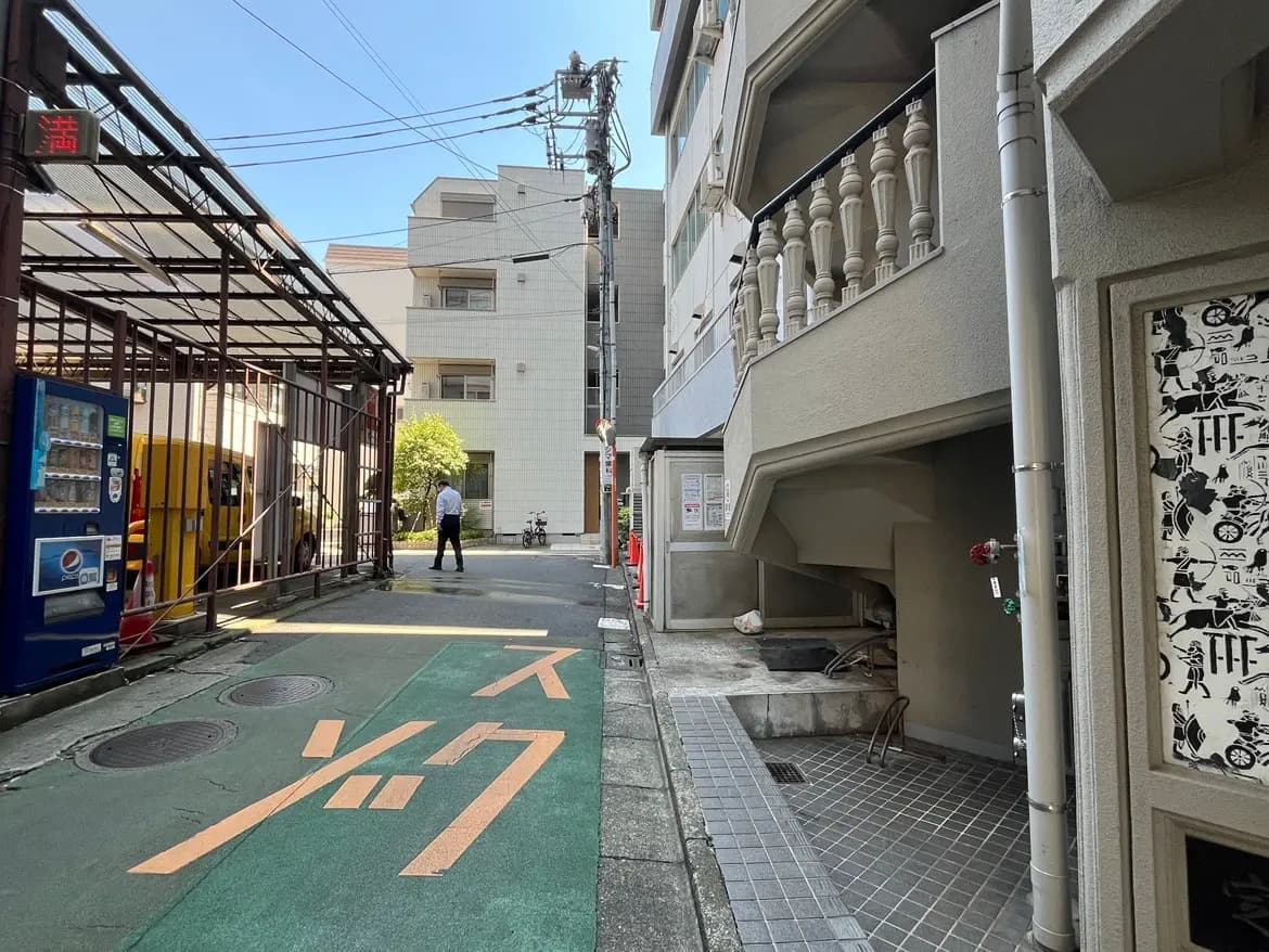 - 住宅街の裏道、両側に建物が立ち並び、自転車と人が通る様子の風景、手前には自販機とコインパーキングの表示が見える。