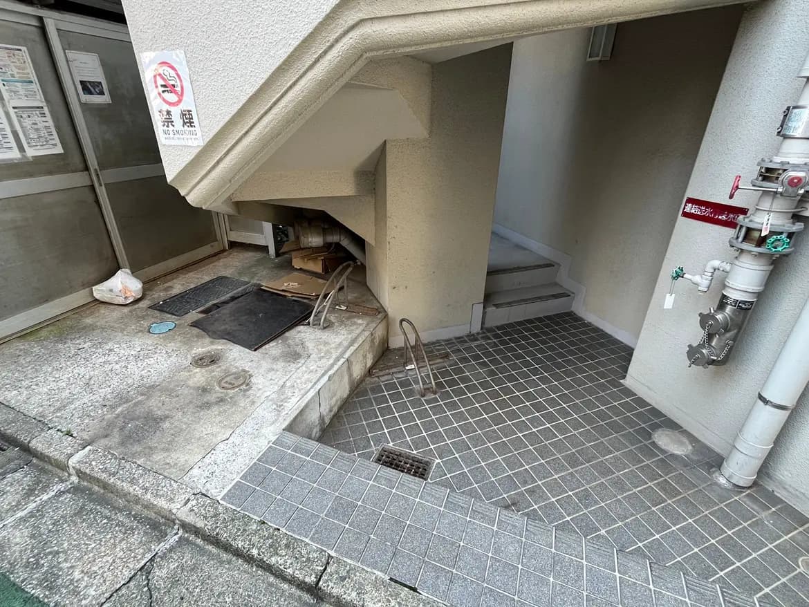 - 建物の裏口付近、階段の下に設置されたスロープ、敷石の床と隣接する配管、禁煙の表示がある場所