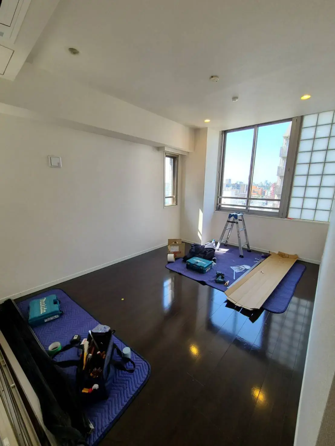 新築住宅の広々とした明るいリビングルーム、光沢のある木製フロア、窓からの自然光、家具の組み立て中