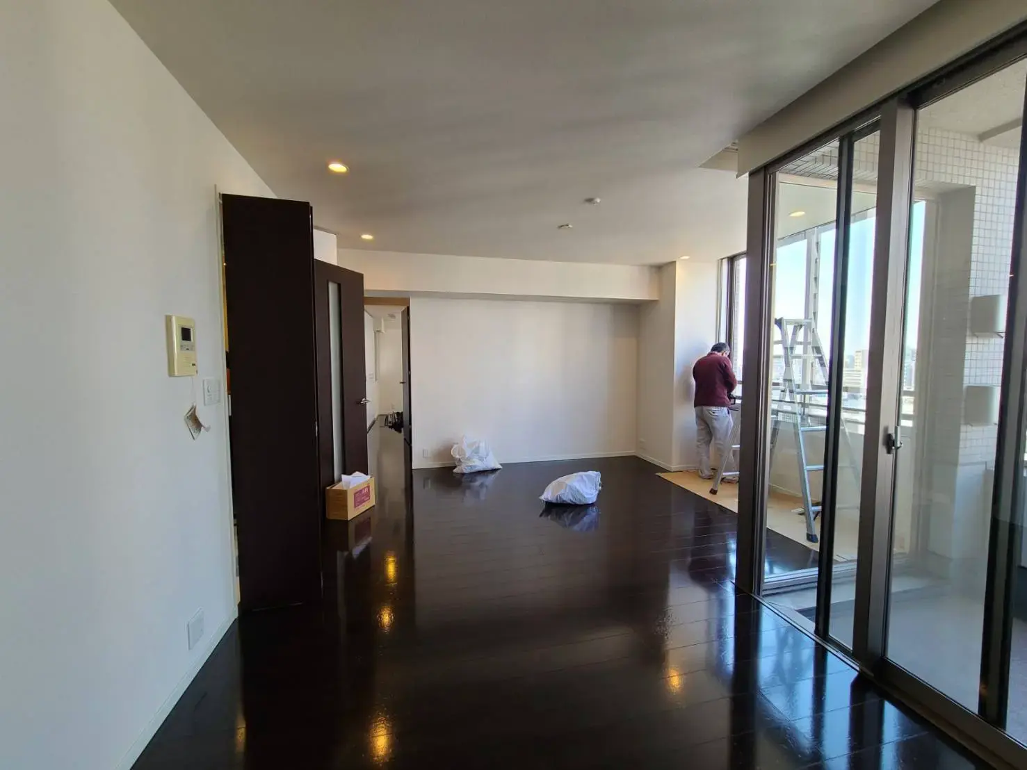 広々とした明るいリビングルーム、光沢のあるダークウッドフロア、ガラスの掃出し窓とバルコニーへのアクセス、部屋の片隅で作業中の人