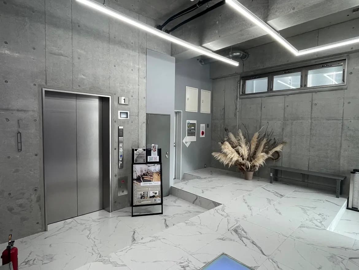 モダンなビルのエントランスホール、コンクリート打ち放しの壁、大理石風タイル床、エレベーターとインフォメーションボード