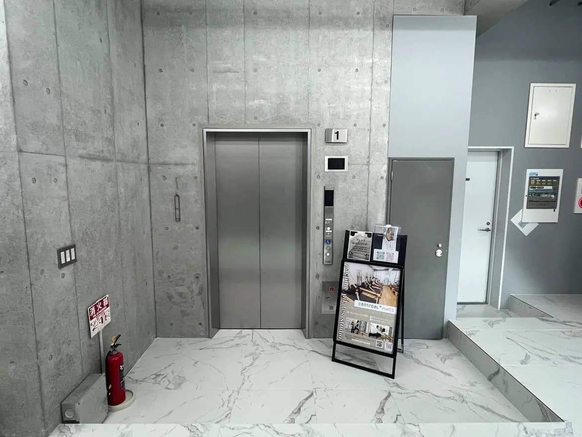 モダンなビルのエントランスホール、コンクリート壁と大理石風タイルの床、エレベーターと消火器が見える